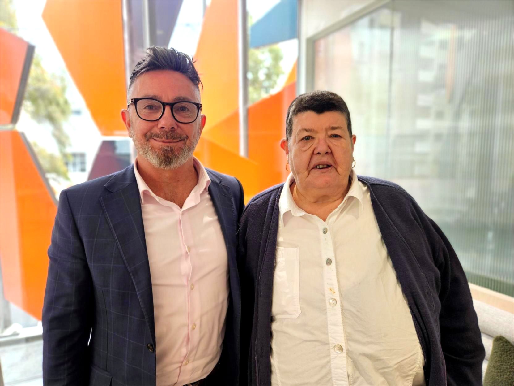 Two people standing close to each other. Sam Hayward is on the left and he is wearing glasses, a blue blazer and pink shirt. Robyn is on the right and she has a white shirt and blue cardigan on. They are both smiling.